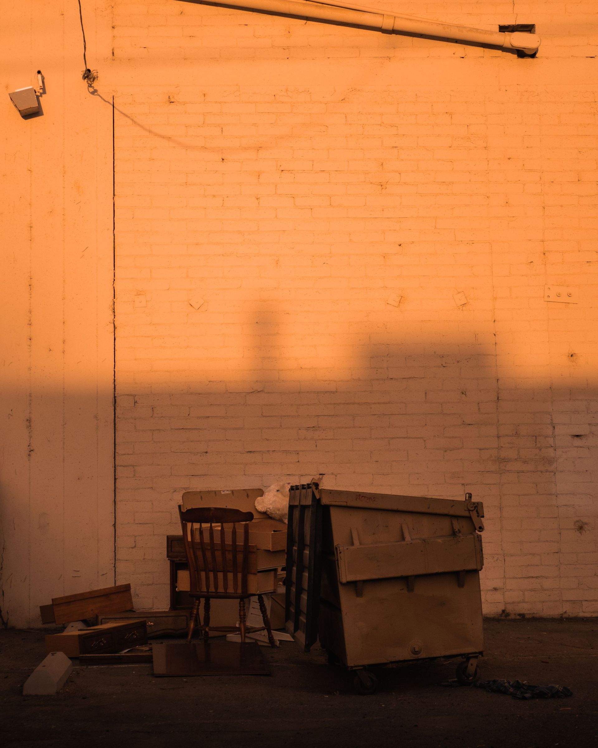 A street scene showing a dumpster and chair against a sunlit wall, depicting urban decay.